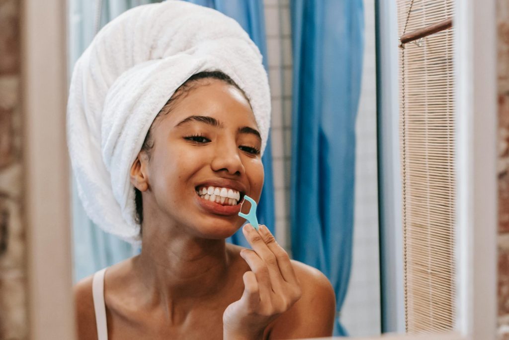 Young woman with towel on head flossing teeth in bathroom mirror, promoting hygiene and wellness.